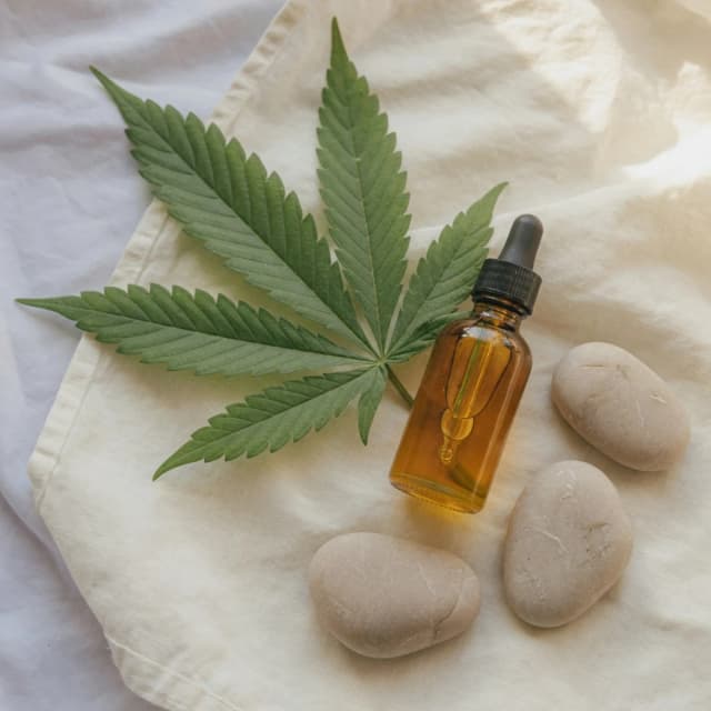 A green hemp leaf, a dropper bottle, and smooth stones on linen — tasteful CBD education still-life