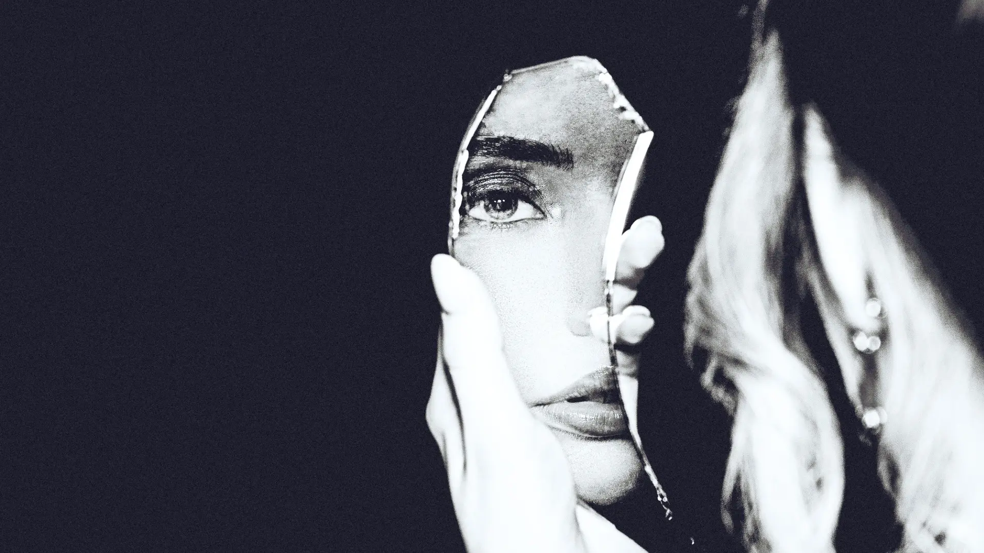 A black and white conceptual photo of a woman holding a mirror fragment that reflects part of her face, representing mirror therapy for chronic pain – Chronic Health Wisdom