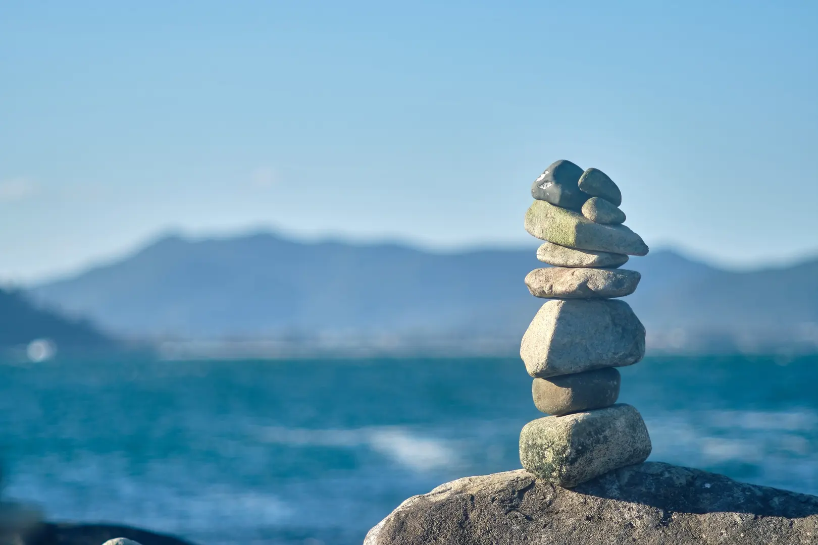Stacked stones balanced on a rock by the ocean with mountains in the distance, symbolizing balance and energy stability through proper diet – Chronic Health Wisdom
