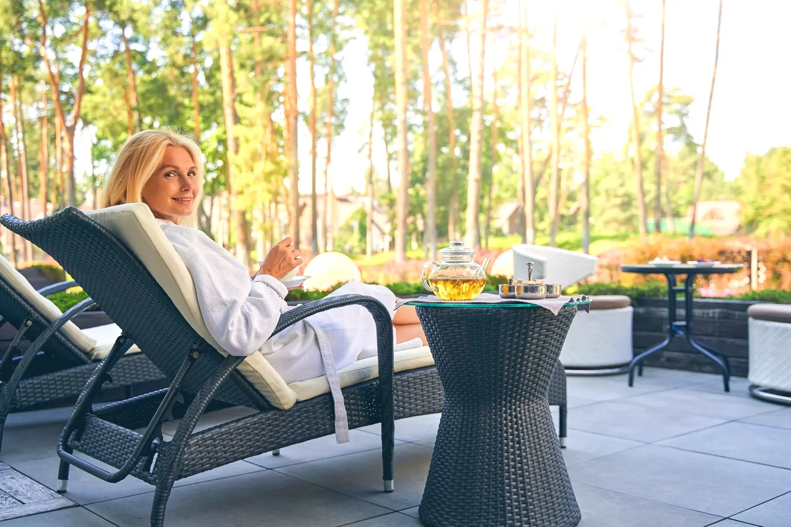 Woman in white robe relaxing on outdoor patio chaise lounge, with tea and books on a side table, promoting restful sleep and relaxation – Chronic Health Wisdom