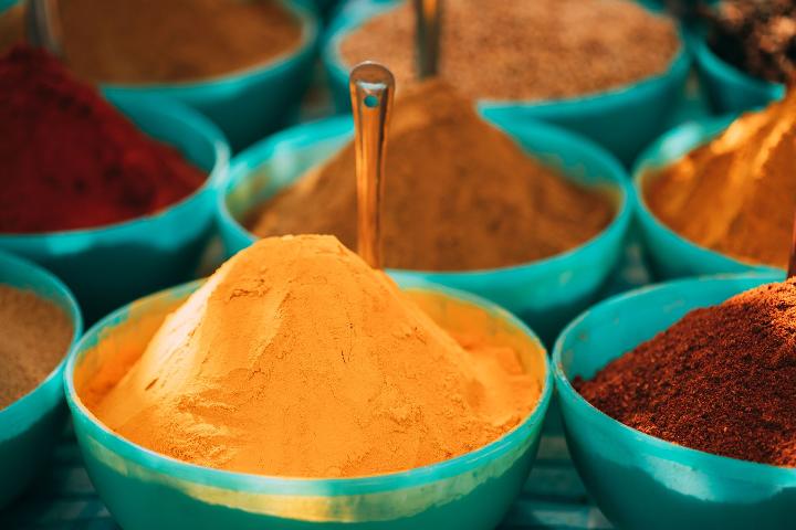 A bowl of tumeric with a spoon in it surrounded by other bowls of spices.