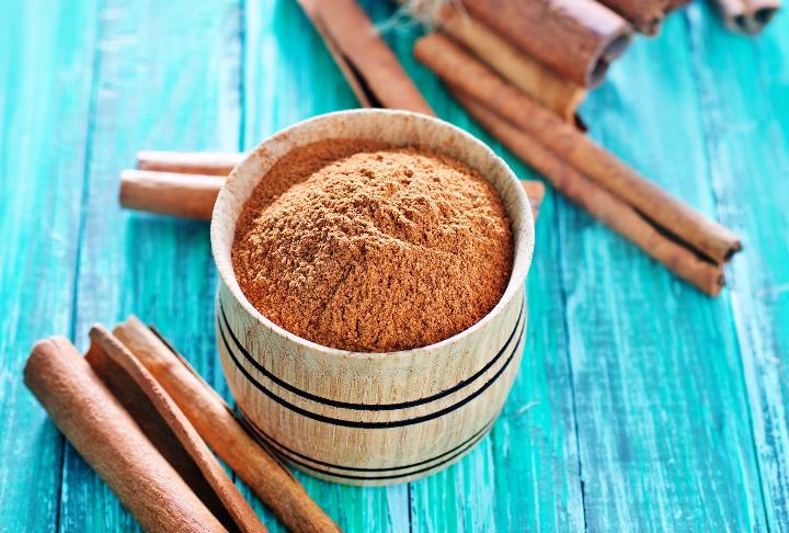Container of cinnamon on a green wooden slab with cinnamon sticks.